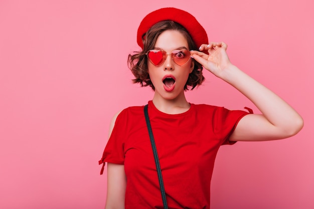 Belle fille en tenue française drôle posant avec une expression de visage surpris. Femme étonnée attrayante aux cheveux ondulés touchant ses lunettes de soleil.