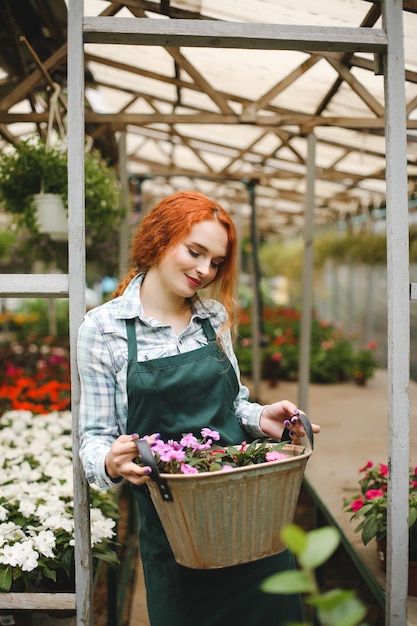 Belle fleuriste rousse en tablier debout avec un panier en métal avec des fleurs dans les mains et fermant rêveusement les yeux