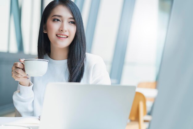 Belle fraîcheur féminine asiatique attrayante sourire joyeux matin travaillant avec ordinateur portable café travail à domicile avec bonheur profiter de la vue sur le jardin jeune femme asiatique avec fenêtre fond de jardin clair