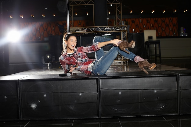 Belle jeune chanteuse pop star souriante avec microphone assis sur la scène dans le club