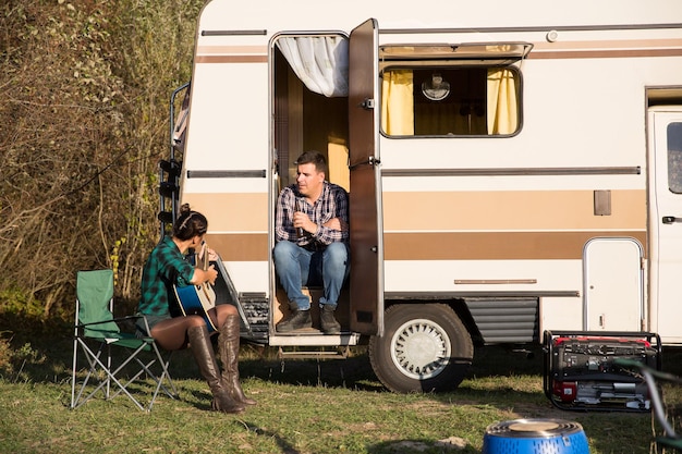 Belle jeune épouse chantant une chanson sur sa guitare pour son mari devant leur camping-car reto dans les montagnes.