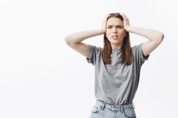 Belle jeune étudiante caucasienne aux cheveux noirs dans des vêtements décontractés, serrant la tête avec les mains, regardant de côté, souffrant de terribles maux de tête après la fête la nuit dernière.