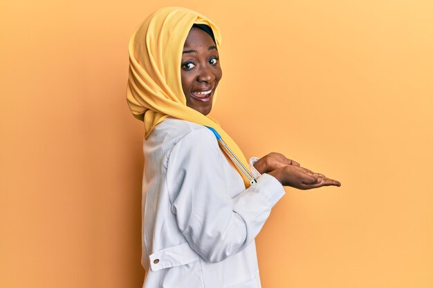 Belle jeune femme africaine portant l'uniforme de médecin et le hijab pointant de côté avec les mains ouvertes paumes montrant l'espace de copie présentant une publicité souriante excitée heureuse