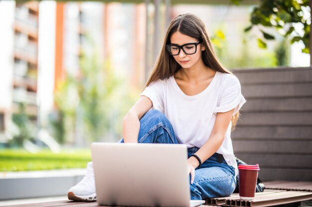 Belle jeune femme à l'aide d'un ordinateur portable assis sur un banc, boire une tasse de café à emporter