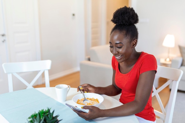 Belle jeune femme américaine arficane textos sur téléphone intelligent à la maison Dame gaie mangeant des flocons de maïs buvant du café du matin et regardant son téléphone portable