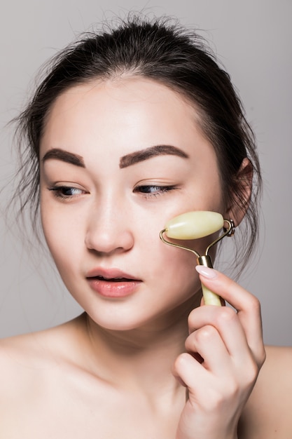 Belle jeune femme asiatique à l'aide d'un rouleau de visage de jade sur sa peau impeccable. Gros plan de visage de beauté. Conceptuel de soins du visage avec des pierres semi-précieuses. isolé sur gris avec espace copie