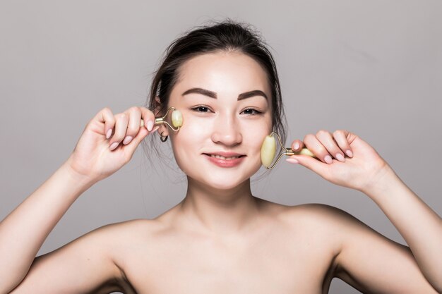 Belle jeune femme asiatique à l'aide d'un rouleau de visage de jade sur sa peau impeccable. Gros plan de visage de beauté. Conceptuel de soins du visage avec des pierres semi-précieuses. isolé sur gris avec espace copie