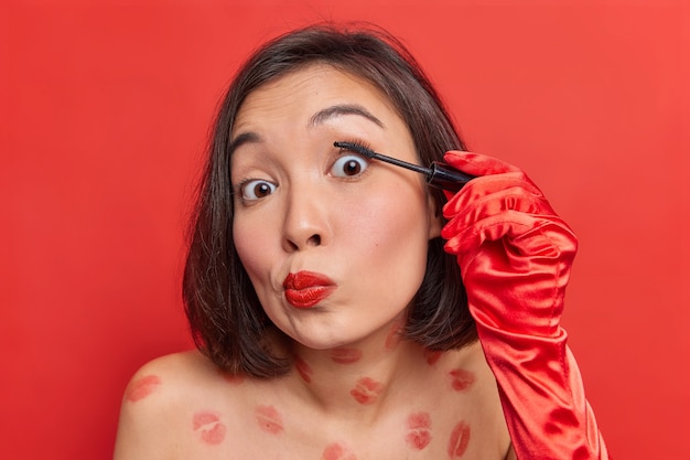 Photo gratuite une belle jeune femme asiatique applique du mascara sur les cils et prépare un maquillage quotidien pour un rendez-vous ou une fête avec un corps nu contre un mur rouge vif
