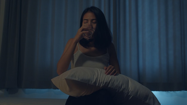 Belle jeune femme asiatique assise sur le lit, prenez un somnifère ou un médicament de nuit dans la chambre. Femme indienne malade en mauvaise santé souffre d'insomnie ou de maux de tête, fille dépressive détient des médicaments antidépresseurs.