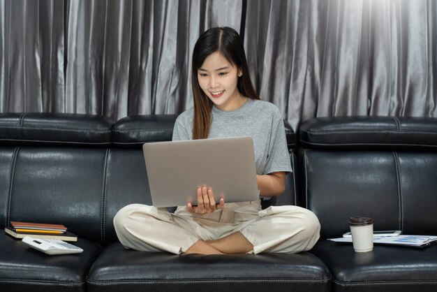 Belle jeune femme asiatique belle travaillant avec un ordinateur portable et un document tout en étant assis au bureau du salon intérieur en tant que pigiste, travaillant en e-coaching, à distance ou travaillant à domicile.