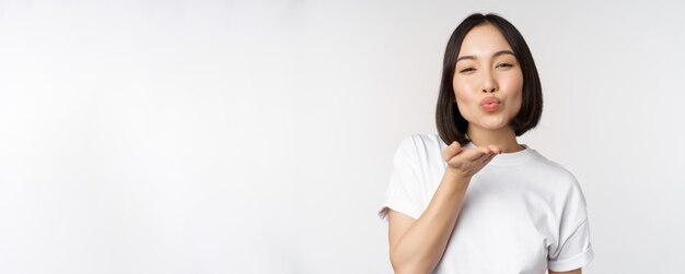 Belle jeune femme asiatique envoyant un baiser aérien et regardant coquette à la caméra debout en t-shirt sur fond blanc