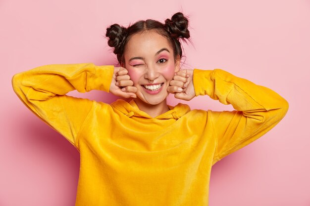 Belle jeune femme asiatique avec l'expression du visage heureux, clignote des yeux et sourit positivement, s'amuse à l'intérieur, deux nœuds de cheveux