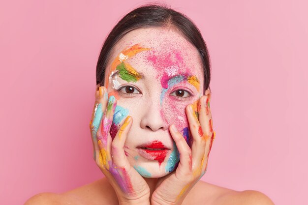 Belle jeune femme asiatique garde les mains sur les joues a de la peinture colorée sur le visage