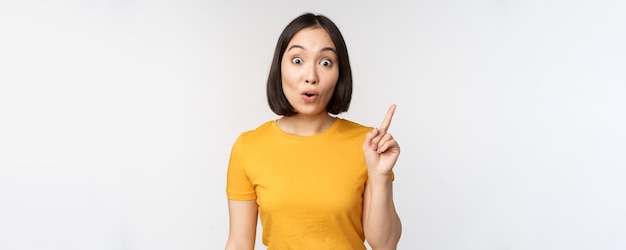 Belle jeune femme asiatique pointant le doigt vers le haut souriant et regardant amusé la caméra montrant l'annonce publicitaire sur le dessus fond blanc