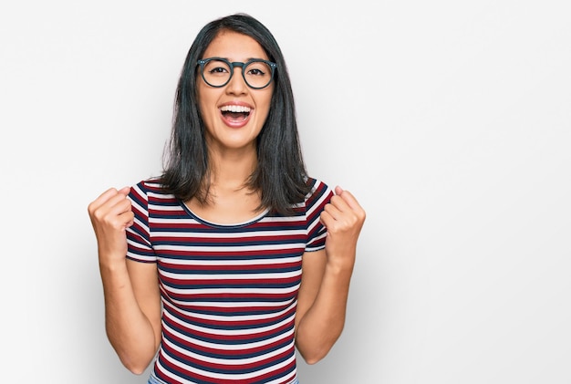 Belle jeune femme asiatique portant des vêtements décontractés et des lunettes célébrant la surprise et l'émerveillement du succès avec les bras levés et les yeux ouverts concept gagnant