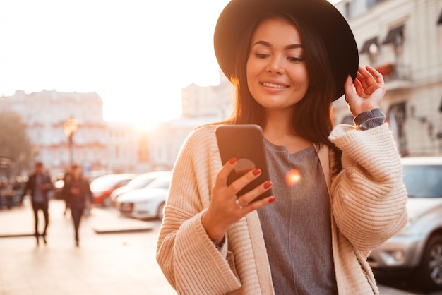 Belle jeune femme asiatique souriante touchant son chapeau tout en vérifiant les nouvelles sur smartphone dans la rue de la ville