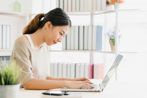 Belle jeune femme asiatique souriante travaillant sur un ordinateur portable sur le bureau dans le salon à la maison