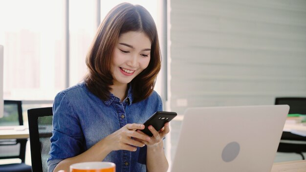 Belle jeune femme asiatique souriante travaillant sur ordinateur portable tout en profitant de l&#39;utilisation de smartphone au bureau.