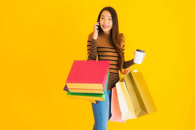 Belle jeune femme asiatique utilise un téléphone mobile intelligent ou un téléphone portable avec une tasse de café et un sac à provisions de couleur sur le mur jaune