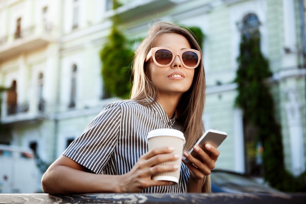 Belle jeune femme assise sur un banc, tenant le café et le téléphone.