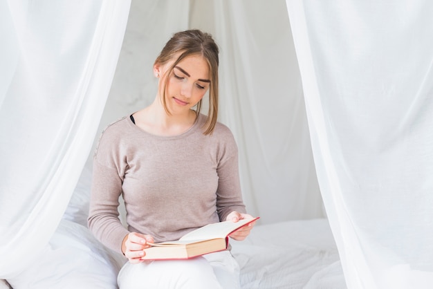Photo gratuite belle jeune femme assise dans son lit, livre de lecture à la maison