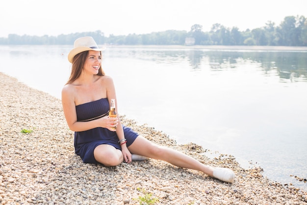 Belle jeune femme assise près de la côte en tenant une bouteille de bière de jus de fruits