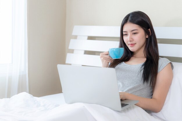 Belle jeune femme assise et tenant une tasse de café et utilise un ordinateur portable pour travailler sur le lit à la maison