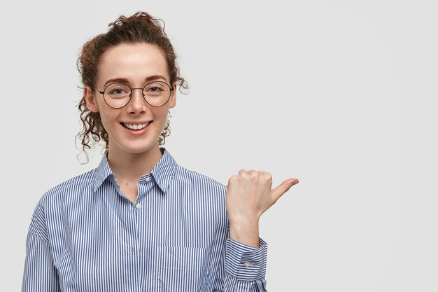 Belle jeune femme attirante de taches de rousseur avec une expression joyeuse, a des taches de rousseur sur la peau, vêtue d'une chemise élégante, pointe de côté avec le pouce