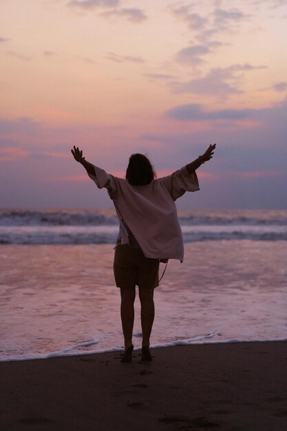 Une belle jeune femme au bord de l'océan au coucher du soleil jouit de la liberté, du lieu, du temps. Vue de dos. vêtements fluides.