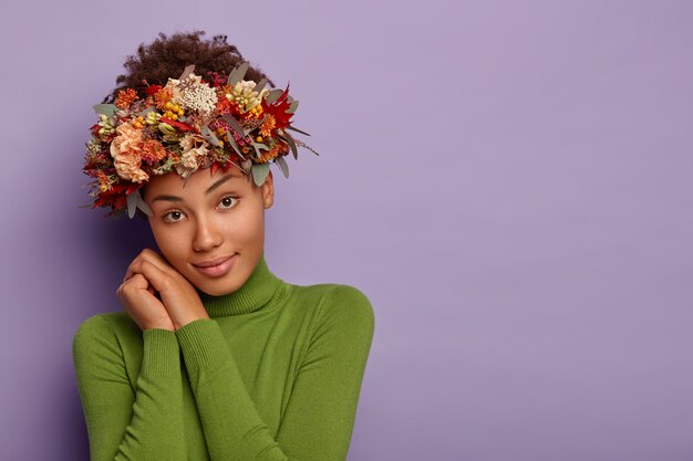 Belle jeune femme d'automne regarde calmement la caméra, se penche des deux mains, porte une couronne de plantes et de feuilles de saison, vêtue de poloneck vert, a une expression faciale détendue, pose à l'intérieur