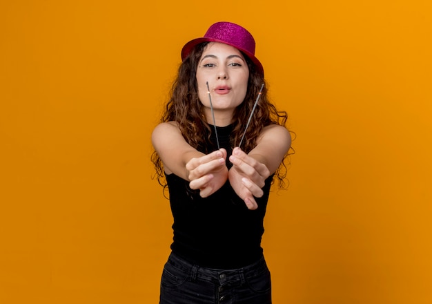 Belle jeune femme aux cheveux bouclés en chapeau de fête tenant des cierges heureux et excité debout sur un mur orange