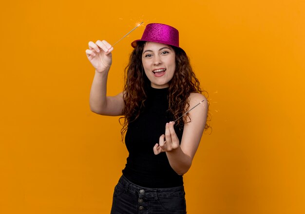 Belle jeune femme aux cheveux bouclés en chapeau de fête tenant des cierges heureux et excité debout sur un mur orange