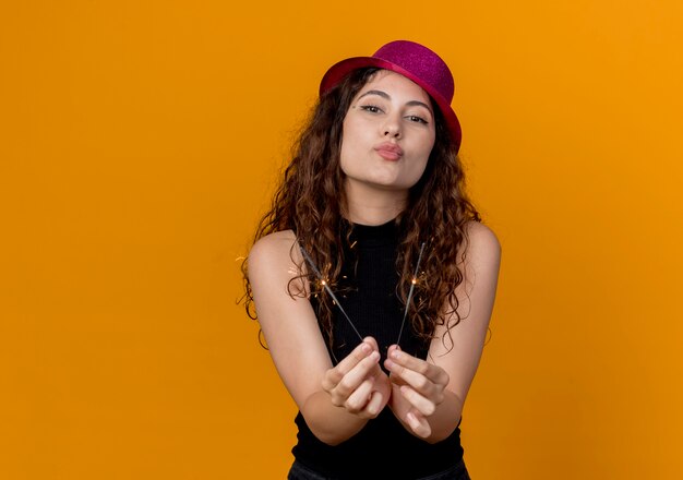 Belle jeune femme aux cheveux bouclés en chapeau de fête tenant des cierges joyeux et joyeux soufflant un baiser debout sur le mur orange