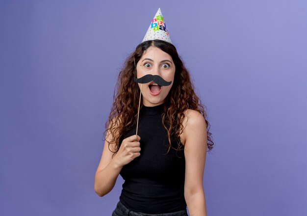 Belle jeune femme aux cheveux bouclés en chapeau de vacances tenant des bâtons de fête moustache heureux et surpris debout sur le mur bleu