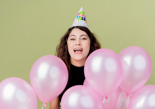 Belle jeune femme aux cheveux bouclés dans un chapeau de vacances avec des ballons à air heureux et excité célébrant la fête d'anniversaire debout sur un mur léger