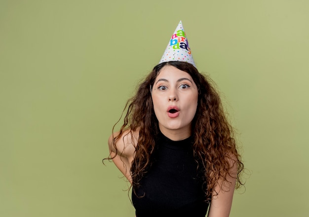 Belle jeune femme aux cheveux bouclés dans un chapeau de vacances concept de fête d'anniversaire surpris debout sur un mur léger