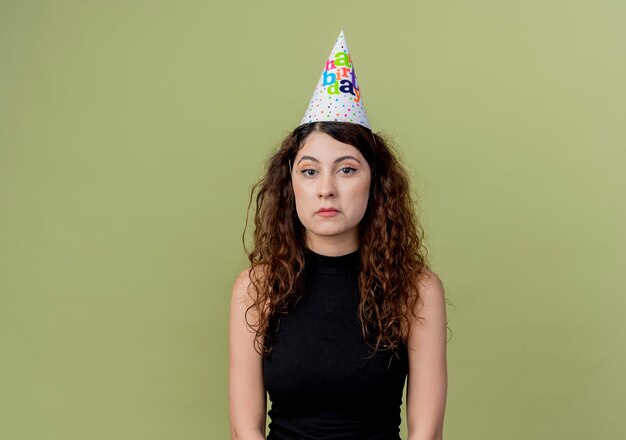 Belle jeune femme aux cheveux bouclés dans un chapeau de vacances avec une expression triste sur le concept de fête d'anniversaire de visage debout sur un mur orange