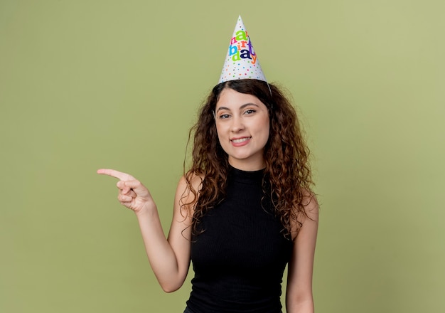 Belle jeune femme aux cheveux bouclés dans un chapeau de vacances pointign avec le doigt sur le côté souriant joyeusement concept de fête d'anniversaire debout sur un mur léger