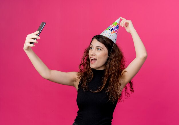 Belle jeune femme aux cheveux bouclés dans un chapeau de vacances prenant selfie souriant joyeusement concept de fête d'anniversaire sur rose