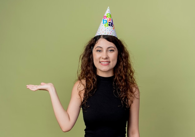 Belle jeune femme aux cheveux bouclés dans un chapeau de vacances présentant avec bras de main souriant joyeusement concept de fête d'anniversaire debout sur un mur léger