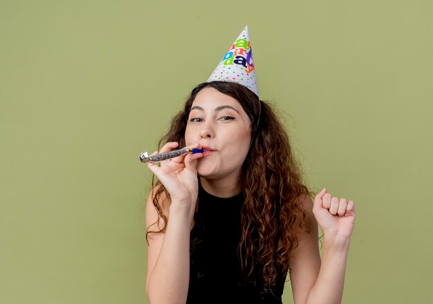 Belle jeune femme aux cheveux bouclés dans un chapeau de vacances sifflet heureux et positif concept de fête d'anniversaire sur la lumière