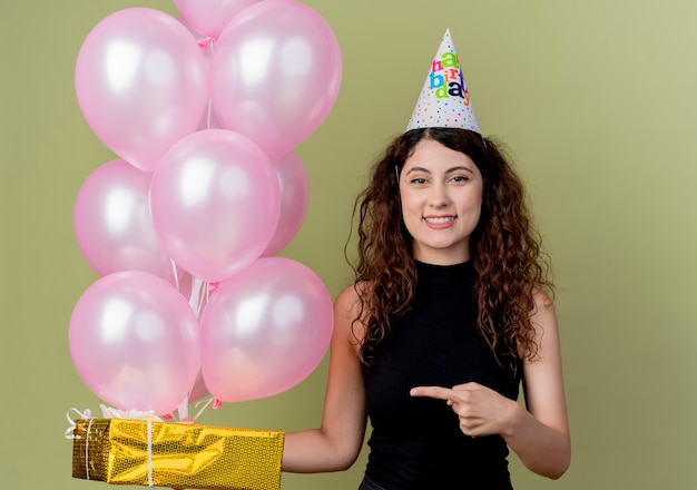 Belle jeune femme aux cheveux bouclés dans un chapeau de vacances tenant des ballons à air et boîte-cadeau pointant avec le doigt sur elle souriant joyeusement heureux et positif debout sur un mur léger