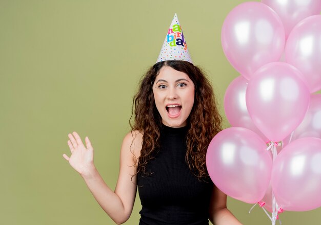 Belle jeune femme aux cheveux bouclés dans un chapeau de vacances tenant des ballons à air comprimé main heureux et excité souriant joyeusement concept de fête d'anniversaire debout sur un mur léger