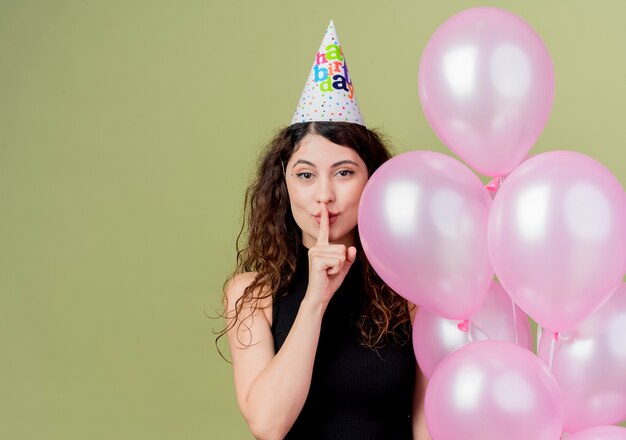 Belle jeune femme aux cheveux bouclés dans un chapeau de vacances tenant des ballons à air faisant le geste de silence avec le doigt sur les lèvres à la notion de fête d'anniversaire confiant debout sur un mur léger