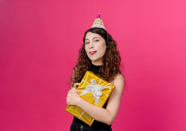 Belle jeune femme aux cheveux bouclés dans un chapeau de vacances tenant boîte-cadeau d'anniversaire souriant joyeusement sticking out tongue concept de fête d'anniversaire debout sur le mur rose