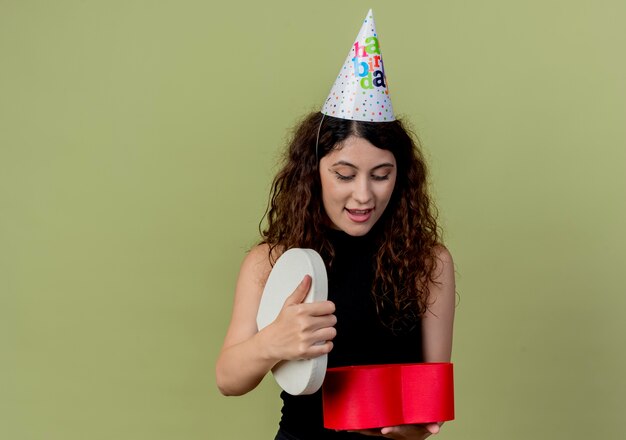 Belle jeune femme aux cheveux bouclés dans un chapeau de vacances tenant une boîte-cadeau en regardant surpris et joyeux anniversaire concept de fête sur la lumière