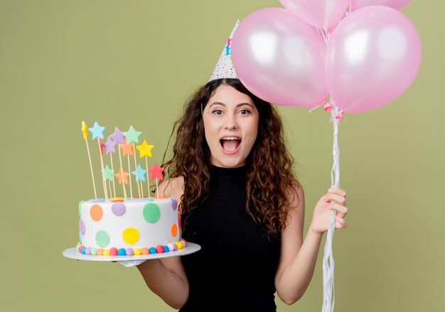 Belle Jeune Femme Aux Cheveux Boucles Dans Un Chapeau De Vacances Tenant Un Gateau D Anniversaire Et Des Ballons A Air Souriant Joyeusement Heureux Et Joyeux Debout Sur Un Mur Leger Photo