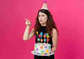 Photo gratuite belle jeune femme aux cheveux bouclés dans un chapeau de vacances tenant le gâteau d'anniversaire pointant du doigt quelque chose à la conception de fête d'anniversaire mécontent debout sur le mur rose
