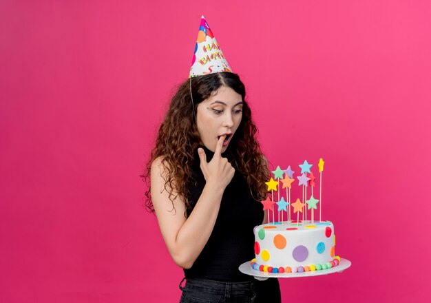 Belle jeune femme aux cheveux bouclés dans un chapeau de vacances tenant le gâteau d'anniversaire en le regardant surpris et étonné concept de fête d'anniversaire debout sur le mur rose