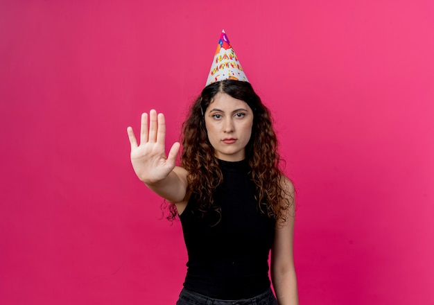 Belle jeune femme aux cheveux bouclés dans un chapeau de vacances avec un visage sérieux faisant signe d'arrêt concept de fête d'anniversaire debout sur un mur rose